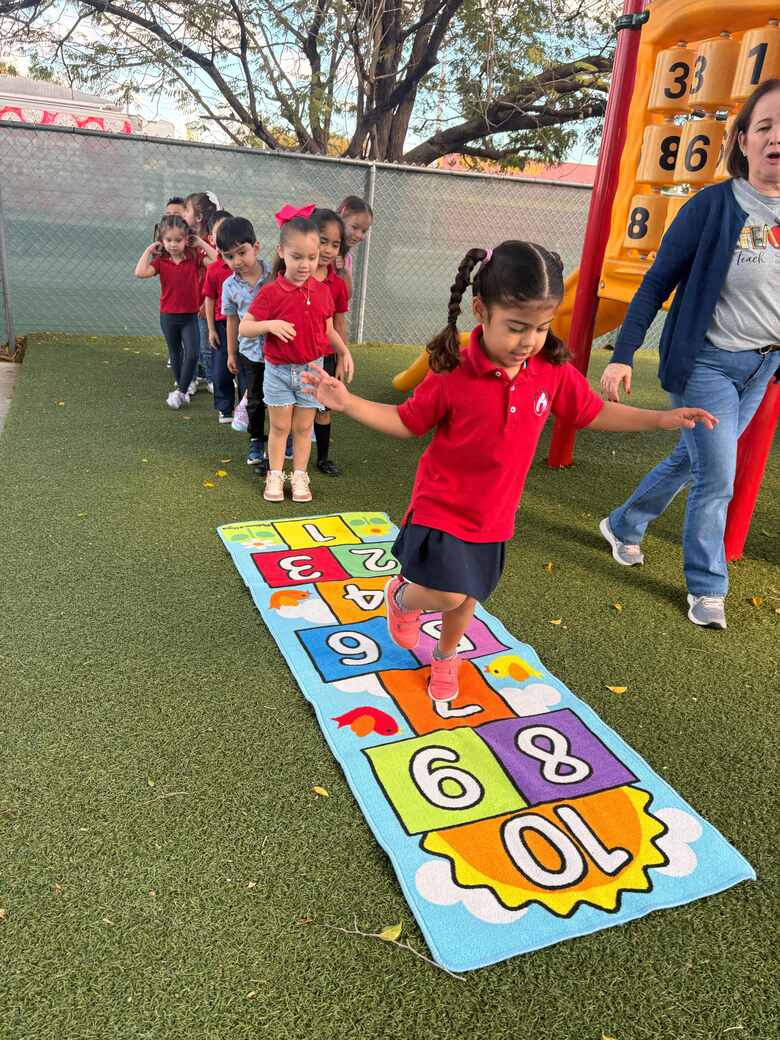 A girl mid-hop along a numbered hopscotch mat on the turf, classmates lined up behind her.