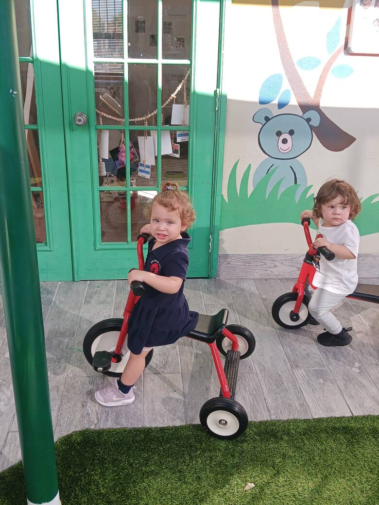 A toddler on a red tricycle in the courtyard, beside a green classroom door with a bear painted on the wall.